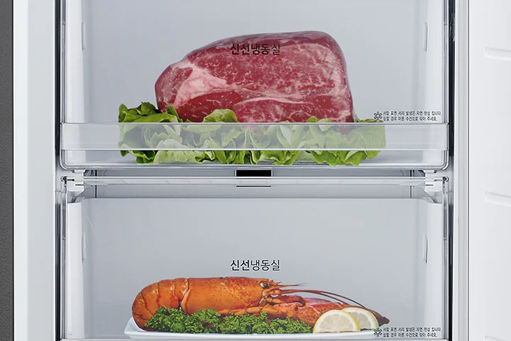 이미지에는 두 개의 투명한 서랍이 있는 냉동고 내부가 보입니다. 상단 서랍에는 신선냉동실이라는 텍스트와 함께 상추 위에 놓인 큰 고기 덩어리가 있습니다. 하단 서랍에는 신선냉동실이라는 텍스트와 함께 접시 위에 놓인 랍스터와 레몬 조각이 보입니다. 서랍의 오른쪽 하단에는 작은 글씨로 ※ 상기 이미지는 실제 제품과 다를 수 있으며, 제품의 성능을 위한 연출된 이미지입니다.라는 문구가 포함되어 있습니다.